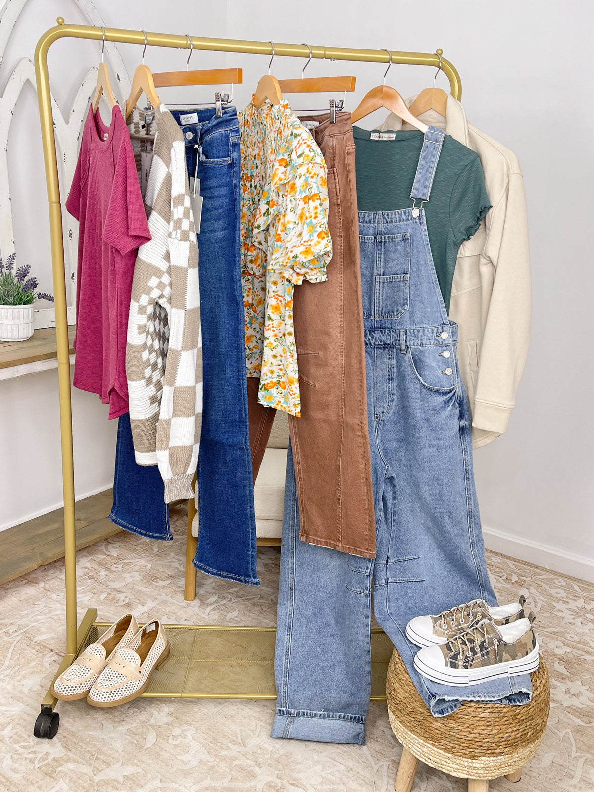 A gold clothing rack styled with multiple outfits, including blue jeans, a brown corduroy poncho over a green shirt, light blue denim overalls, a floral top, a checkered cardigan, a pink t-shirt, and a cream jacket. A pair of woven slip-on shoes sits on the floor next to a small woven stool holding folded jeans and patterned sneakers.