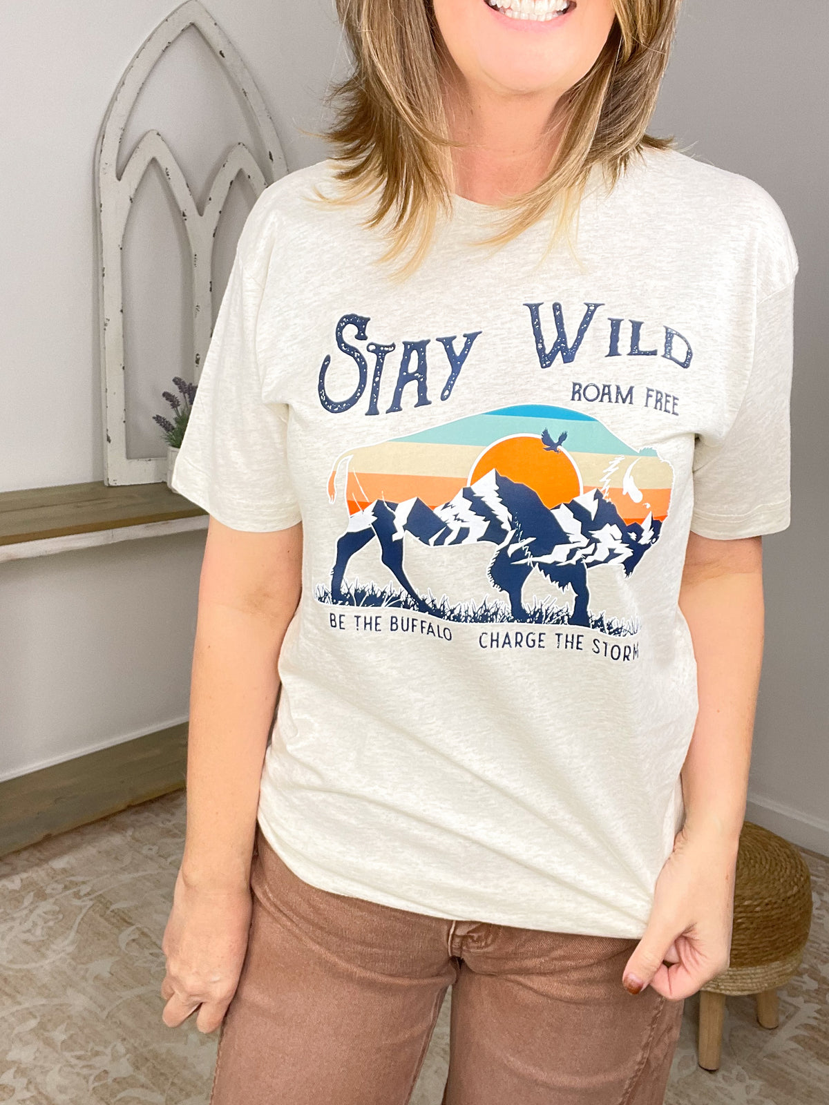 A woman wearing a light heather gray graphic t-shirt with a blue, orange, and yellow mountain sunset design and the text 'Stay Wild', 'Roam Free', and 'Be the Buffalo Charge the Mountain'.