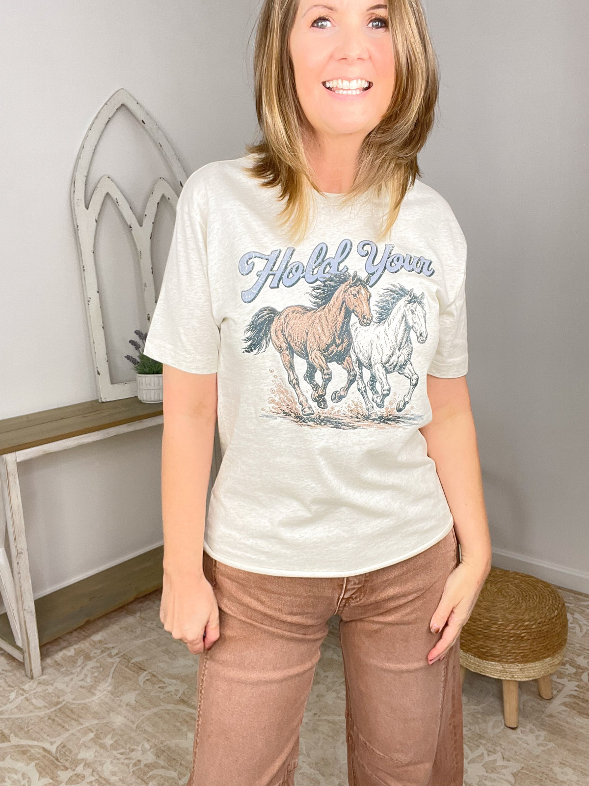 A woman wearing a cream-colored graphic t-shirt with a horse design and the text 'Hold Your', paired with brown pants, standing in a home interior setting.